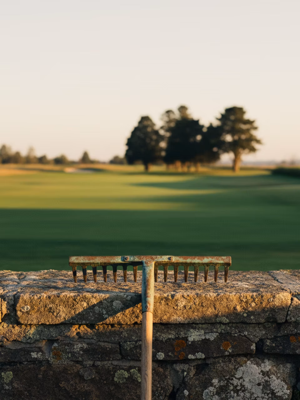 A weathered brass bunker rake leaning on a low stone wall at golden hour